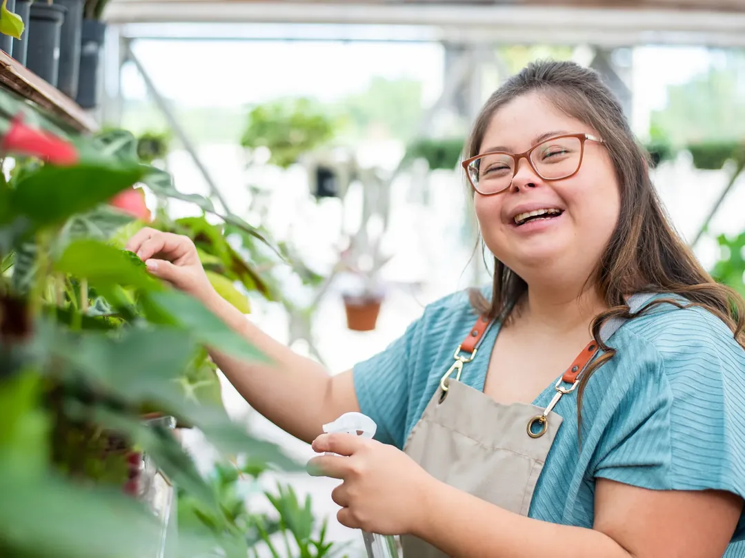 A young woman in an apron tending to plants in a nursery, laughing.